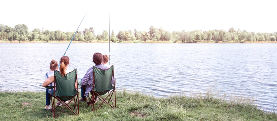 Great view of big river flowing. There is a family sitting by its shore and enjoying the moment. They are holding kids on the lap. Children are holding fish rods in hands. Outdoor Activities